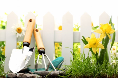 A close-up view of a small garden area in an outdoor space, featuring two gardening tools—a trowel and a hand rake—with wooden handles resting against a white picket fence. The garden bed in front of the fence is filled with lush, green grass and vibrant yellow daffodil flowers in bloom, alongside a few white daisies. Behind the fence, blurred foliage suggests a sunny day with natural light illuminating the scene. The surface of the soil in the garden bed appears moist and well-maintained, contributing to a healthy and tidy appearance suitable for gardening services offered by Lawn Mowing Wembley in the Wembley area. The overall composition highlights a neat and cared-for outdoor environment, ideal for lawn and garden maintenance, with clear focus on the tools and flowering plants that define a vibrant, inviting garden space.